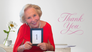 Rita Wilkins smiling in her red dress holding a small framed card with the number one, seated at a table with a white daisy in a glass vase beside her. The words “Thank You” appear in soft pink script on a light background.