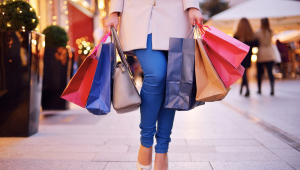 Person walking outdoors holding multiple colorful shopping bags in both hands, suggesting excess shopping and consumer overwhelm.