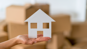 Hand holding a white paper cutout of a house with moving boxes blurred in the background, symbolizing downsizing, relocation, or transitioning to a smaller home... downsizing.