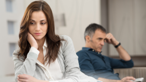 Woman reflecting during gray divorce while partner sits in background, symbolizing emotional distance and loss of shared home and life after divorce over 50.