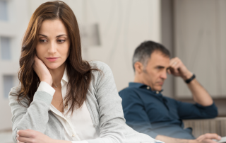Woman reflecting during gray divorce while partner sits in background, symbolizing emotional distance and loss of shared home and life after divorce over 50.