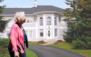 Older woman standing outside a large suburban home, looking thoughtfully at the house, symbolizing reflection about whether a big home still fits life after 60 and the idea of downsizing or right-sizing later in life.