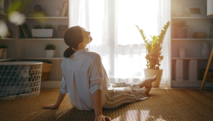 Woman sitting peacefully on the floor in a bright, clutter-free room, surrounded by natural light, plants, and simple home décor, symbolizing calm, intentional living, and reclaiming your space.