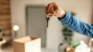 Close-up of a hand holding a set of house keys in front of a softly blurred interior space with a moving box and minimal furnishings, symbolizing transition, downsizing, and a new beginning.