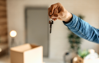 Close-up of a hand holding a set of house keys in front of a softly blurred interior space with a moving box and minimal furnishings, symbolizing transition, downsizing, and a new beginning.