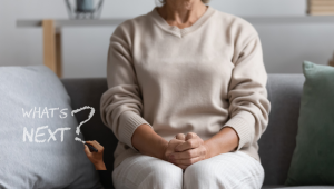 Woman in her 60s sitting thoughtfully on a sofa with hands clasped, beside the words “What’s Next?” written on a soft, blurred background, representing reflection, life transitions, downsizing, and deciding what comes next after 60.