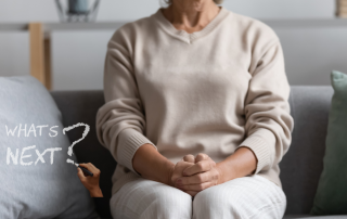 Woman in her 60s sitting thoughtfully on a sofa with hands clasped, beside the words “What’s Next?” written on a soft, blurred background, representing reflection, life transitions, downsizing, and deciding what comes next after 60.
