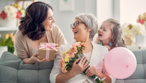 Three generations of women smile together on a couch as a mother receives flowers and a gift for Mother’s Day, capturing love, connection, and appreciation.