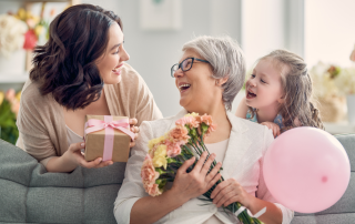 Three generations of women smile together on a couch as a mother receives flowers and a gift for Mother’s Day, capturing love, connection, and appreciation.