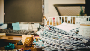 A large stack of paperwork and documents sits on a cluttered home office desk beside a mug, computer, keyboard, calendar, and office supplies, representing paperwork stress and paper clutter.