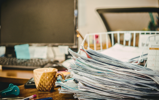 A large stack of paperwork and documents sits on a cluttered home office desk beside a mug, computer, keyboard, calendar, and office supplies, representing paperwork stress and paper clutter.