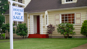House for sale sign in front of a traditional family home, representing the emotional decision many baby boomers face when considering selling or downsizing.