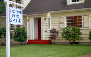 House for sale sign in front of a traditional family home, representing the emotional decision many baby boomers face when considering selling or downsizing.
