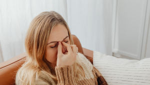 Woman sitting on a couch with her eyes closed, gently pinching the bridge of her nose, looking overwhelmed and stressed while decluttering.